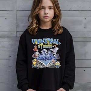 Op de afbeelding: Zwarte sweatshirt met de tekst "UNIVERSAL STUDIOS" in blauw en geel. De afbeelding bevat illustraties van een kasteel, Minions en andere themaparkelementen. De sweatshirt wordt door een persoon gedragen.