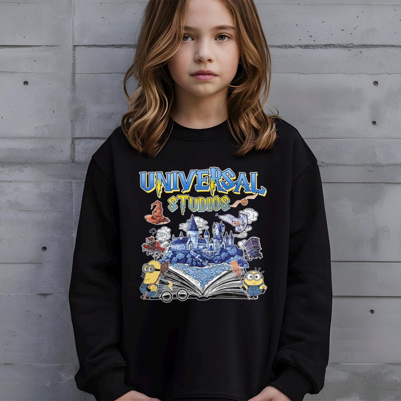 Op de afbeelding: Zwarte sweatshirt met de tekst "UNIVERSAL STUDIOS" in blauw en geel. De afbeelding bevat illustraties van een kasteel, Minions en andere themaparkelementen. De sweatshirt wordt door een persoon gedragen.