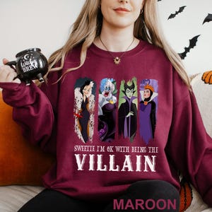 Puede incluir: Sudadera granate con cuatro personajes de villanos de dibujos animados. El texto "Sweetie I'm OK with being the Villain" est&aacute; impreso en blanco debajo de los personajes. La palabra "MAROON" est&aacute; impresa en la parte inferior de la sudadera.