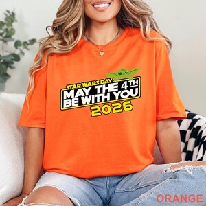 May include: Orange t-shirt with the text "STAR WARS DAY MAY THE 4TH BE WITH YOU 2026" in a black and yellow rectangular design. A green cartoon character is above the text. The shirt is a solid orange color.