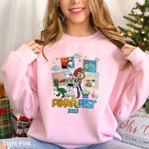 Może przedstawiać: Jasnor&oacute;żowa bluza z kolorową grafiką postaci z film&oacute;w Pixar, w tym Buzzem Astralem i Woodym. Projekt zawiera napis "PIXAR FEST 2025". Bluza ma okrągły dekolt.