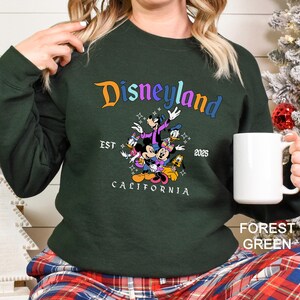 Puede incluir: Sudadera verde bosque con un gr&aacute;fico de Disneyland. El dise&ntilde;o presenta a Mickey Mouse, Minnie Mouse, el Pato Donald, Goofy y Pluto. El texto dice "Disneyland California EST 2025". Se sostiene una taza blanca en la mano.