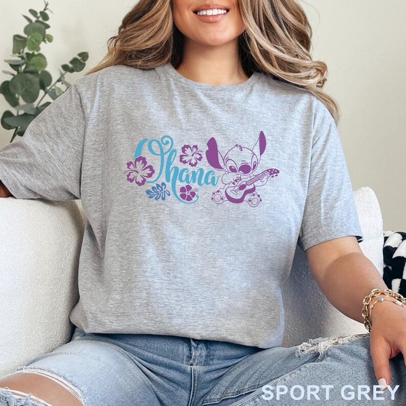 Op de afbeelding: Een sportgrijs t-shirt met een grafische print van het personage Stitch die ukelele speelt naast het woord Ohana in blauw schrift, omringd door paarse hibiscusbloemen en tropische bladeren, met de tekst SPORT GREY in de onderste hoek.