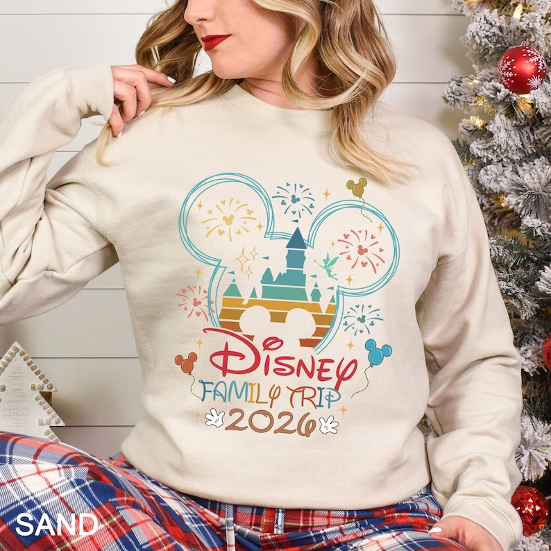 May include: A sand-colored sweatshirt with a colorful Disney Family Trip 2026 graphic. The design features a castle, fireworks, and Mickey Mouse ears. The sweatshirt has long sleeves.