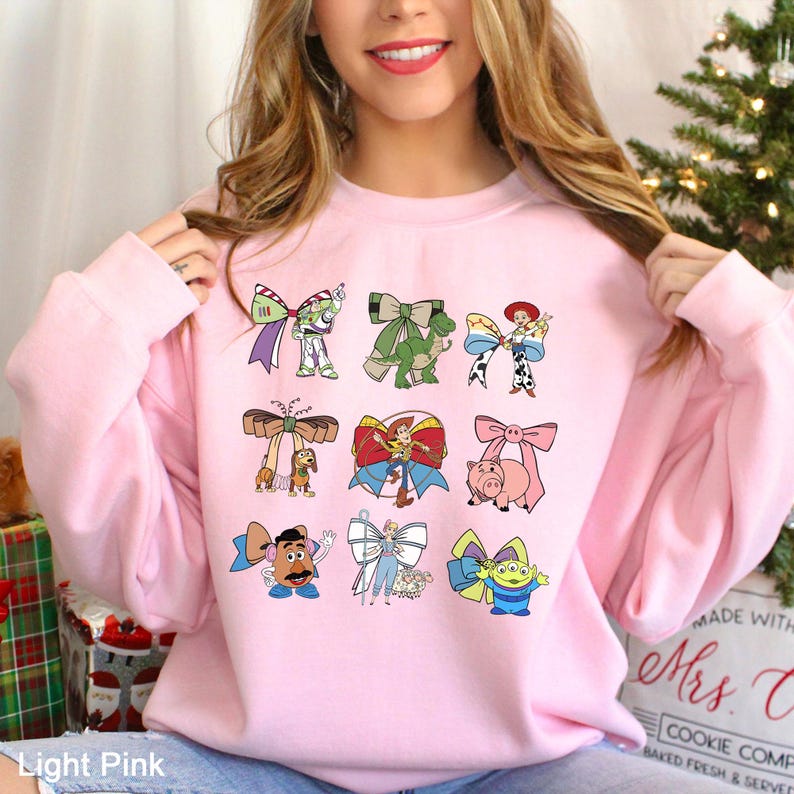 May include: Light pink sweatshirt featuring colorful bow designs with cartoon characters. The bows have characters like Buzz Lightyear, Rex, Woody, and others. The text "Light Pink" is at the bottom.