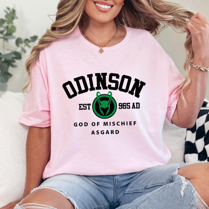 May include: A light pink t-shirt with the word "ODINSON" in black block letters. Below the word is a green and black graphic and the text "EST 965 AD". The shirt also has the text "GOD OF MISCHIEF ASGARD".