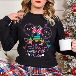 May include: Black sweatshirt with a colorful Disney Family Trip 2026 graphic. The design features a Minnie Mouse bow, fireworks, and a castle silhouette. The text "Disney Family Trip 2026" is in a playful font.