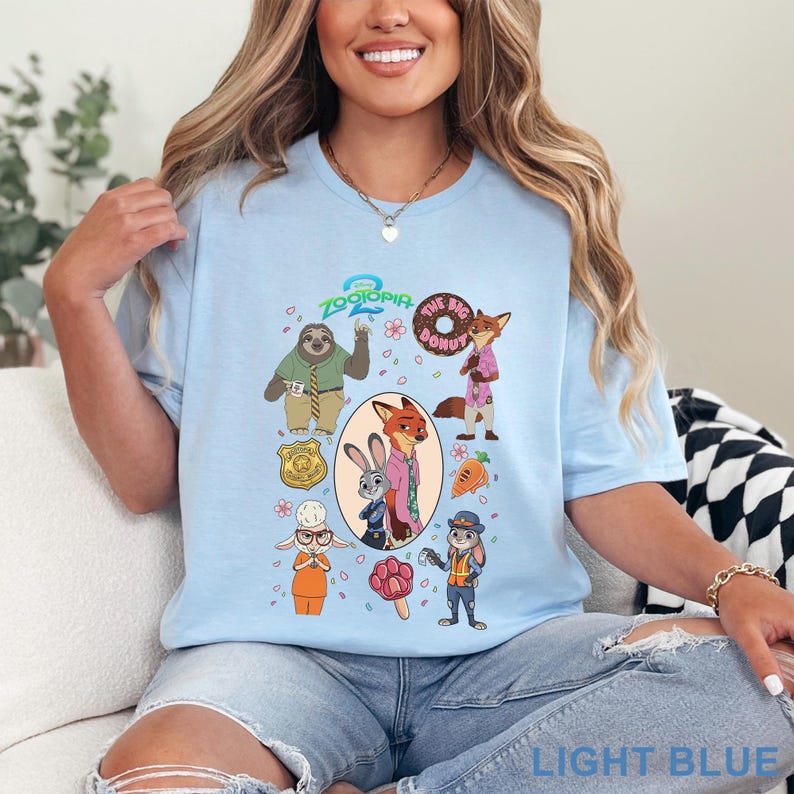 K&ouml;nnte beinhalten: Ein hellblaues T-Shirt mit einem Grafikdruck der Charaktere aus Zoomania, darunter Judy Hopps, Nick Wilde, das Faultier Flash und Vizeb&uuml;rgermeisterin Bellwether, sowie Symbole wie ein Donut, eine Karotte und ein pfotenf&ouml;rmiges Eis am Stiel.