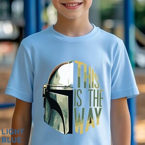 Pu&ograve; includere: Una t-shirt a maniche corte color azzurro con una stampa grafica di un casco metallico sul lato sinistro e il testo "THIS IS THE WAY" in lettere maiuscole gialle e verdi sul lato destro, centrato sul petto del capo di abbigliamento.