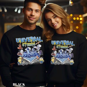 Op de afbeelding: Zwarte sweatshirts met een kleurrijke afbeelding met de woorden "UNIVERSAL STUDIOS" in blauw en geel. De afbeelding bevat een kasteel, Minions en andere cartoon elementen. Het woord "BLACK" staat onderaan gedrukt.