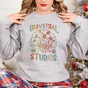 Pode incluir: Camisola cinza desportiva com um gr&aacute;fico colorido da Universal Studios. O design inclui as palavras "UNIVERSAL STUDIOS" e v&aacute;rias imagens e personagens do parque tem&aacute;tico. A camisola &eacute; de gola redonda e manga comprida.