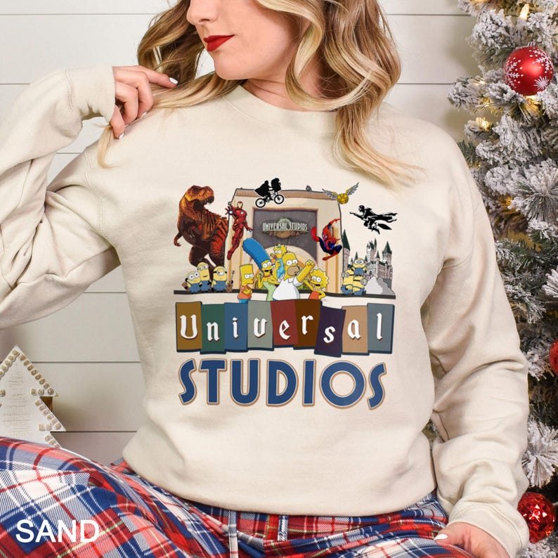 May include: A sand-colored sweatshirt featuring a colorful graphic of Universal Studios characters and the park's logo. The design includes dinosaurs, superheroes, and cartoon characters. The text "Universal Studios" is displayed prominently.