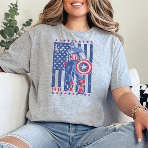 May include: A gray t-shirt featuring a graphic of Captain America with his shield, standing in front of a distressed American flag. The year "1941" is printed below the graphic. The design is in red, white, and blue.
