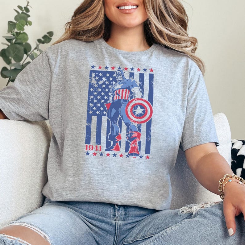 May include: A gray t-shirt featuring a graphic of Captain America with his shield, standing in front of a distressed American flag. The year "1941" is printed below the graphic. The design is in red, white, and blue.