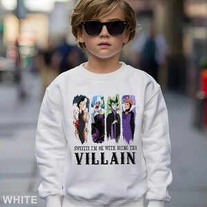 Puede incluir: Sudadera blanca con un dise&ntilde;o de cuatro villanos de dibujos animados: Cruella de Vil, &Uacute;rsula, Mal&eacute;fica y la Reina Malvada. El texto en la sudadera dice "Sweetie I'm ok with being the Villain". La sudadera la lleva una persona con gafas de sol.