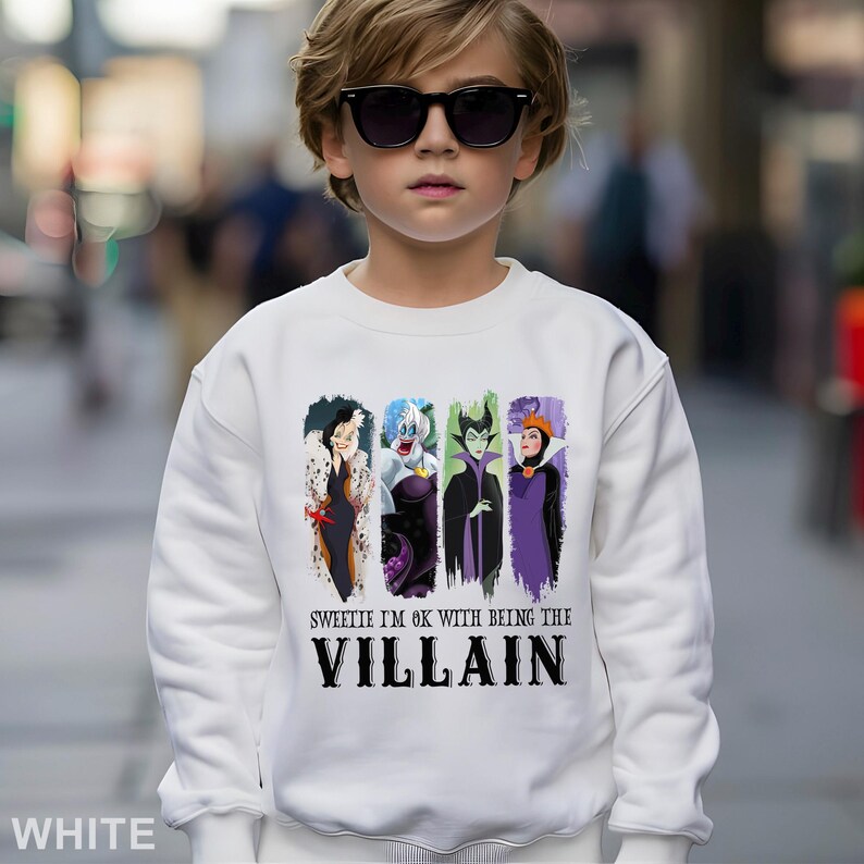 Puede incluir: Sudadera blanca con un dise&ntilde;o de cuatro villanos de dibujos animados: Cruella de Vil, &Uacute;rsula, Mal&eacute;fica y la Reina Malvada. El texto en la sudadera dice "Sweetie I'm ok with being the Villain". La sudadera la lleva una persona con gafas de sol.
