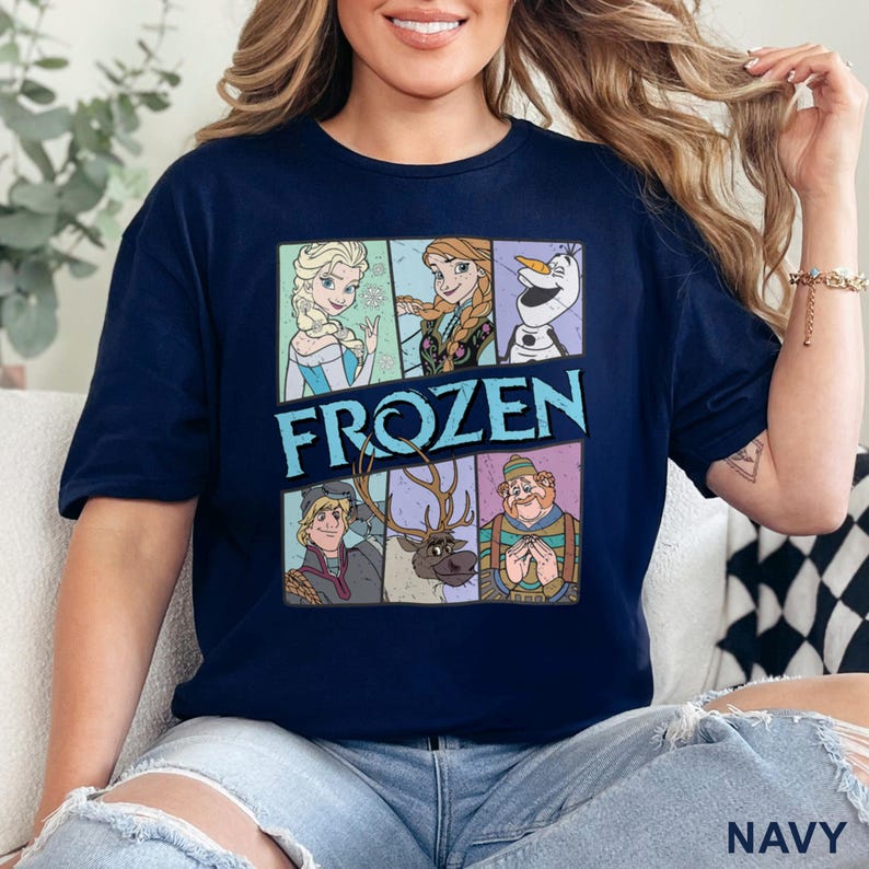 May include: Navy blue t-shirt featuring a colorful graphic of characters from the movie "Frozen." The design includes Elsa, Anna, Olaf, Kristoff, Sven, and a character in a hat. The word "FROZEN" is displayed in large, blue letters.