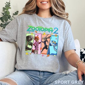 May include: Sport grey t-shirt with the text "ZOOTOPIA 2" and character illustrations from the film. The characters are in rectangular frames with floral and star accents. The shirt is a crew neck.