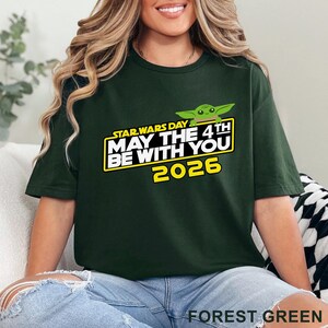 May include: Forest green t-shirt featuring the text "STAR WARS DAY MAY THE 4TH BE WITH YOU 2026" in white and yellow, with a green cartoon character. The shirt is a solid color.
