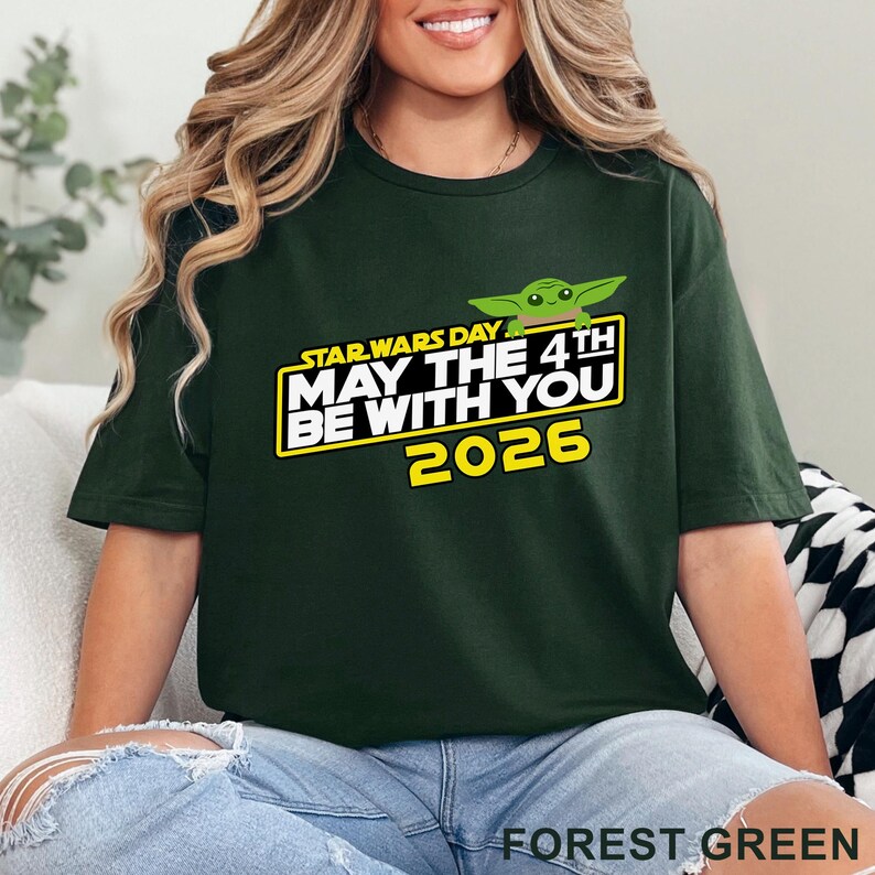 May include: Forest green t-shirt featuring the text "STAR WARS DAY MAY THE 4TH BE WITH YOU 2026" in white and yellow, with a green cartoon character. The shirt is a solid color.