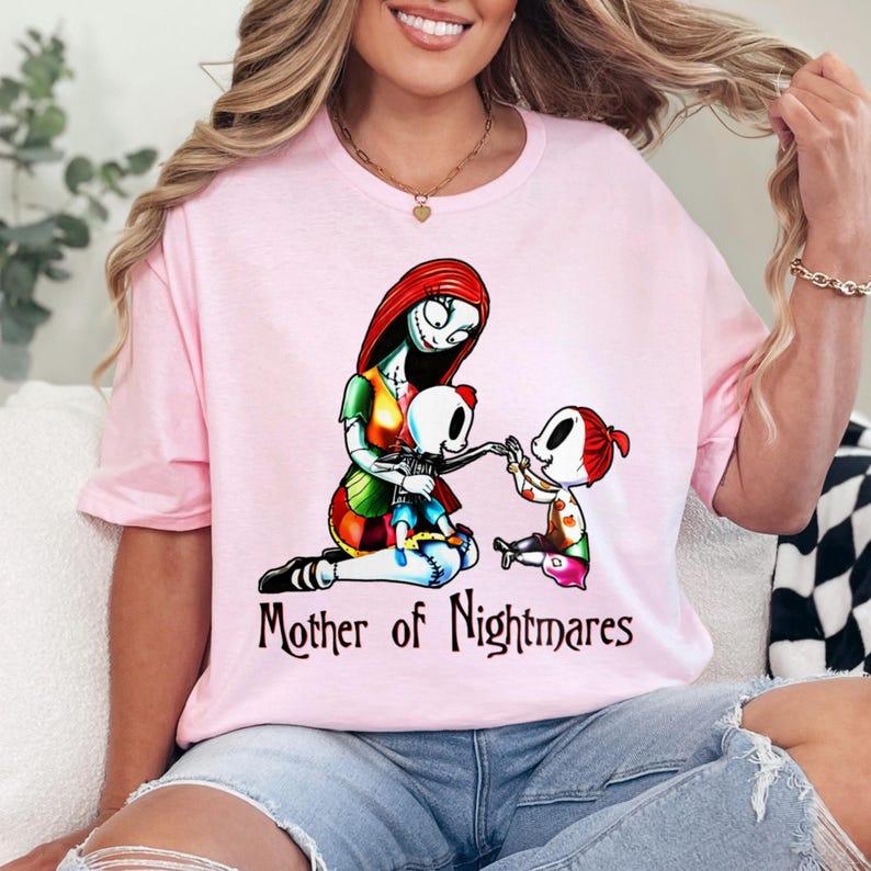 Puede incluir: Camiseta rosa claro con un gr&aacute;fico de un personaje con cabello rojo y apariencia cosida, con dos personajes m&aacute;s peque&ntilde;os. El texto "Mother of Nightmares" est&aacute; impreso debajo. La camiseta es de material suave.