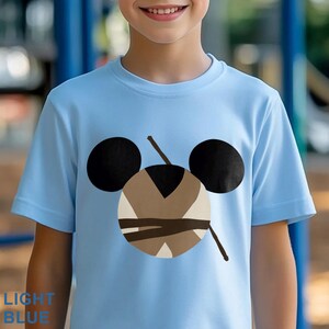 May include: Light blue t-shirt featuring a cartoon design with black circles and a brown and beige face. The shirt has short sleeves and a crew neck. The text "LIGHT BLUE" is at the bottom left of the image.