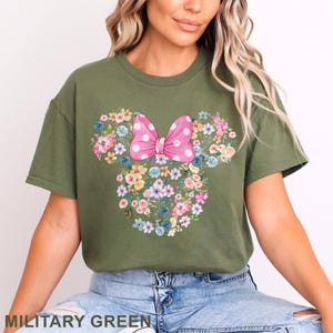 May include: Military green t-shirt featuring a floral Minnie Mouse design. The design includes a pink polka dot bow and a floral arrangement in the shape of Minnie's head. The text "MILITARY GREEN" is at the bottom.