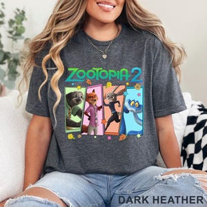 May include: Dark grey t-shirt with the text "ZOOTOPIA 2" and a colourful graphic of characters from the animated film. The graphic includes a sloth, fox, rabbit, and snake. The words "DARK HEATHER" are printed at the bottom.