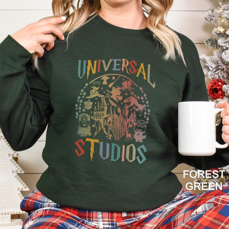 Pode incluir: Camisola verde floresta com um gr&aacute;fico colorido "UNIVERSAL STUDIOS" com personagens de parques tem&aacute;ticos. A camisola &eacute; de gola redonda e tem mangas compridas. Uma caneca branca &eacute; segurada na m&atilde;o.