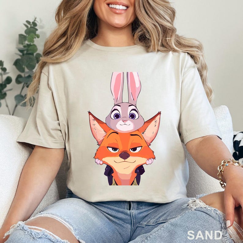 May include: Sand-colored t-shirt featuring a cartoon illustration of a fox and a rabbit. The fox is orange and the rabbit is gray with pink ears. The word "SAND" is printed at the bottom right.