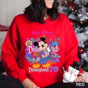 May include: A red sweatshirt featuring a colorful graphic of Mickey Mouse in front of Disneyland Resort imagery. The design includes the text "Walt Disney" and "Disneyland 70". The sweatshirt is a vibrant red color.