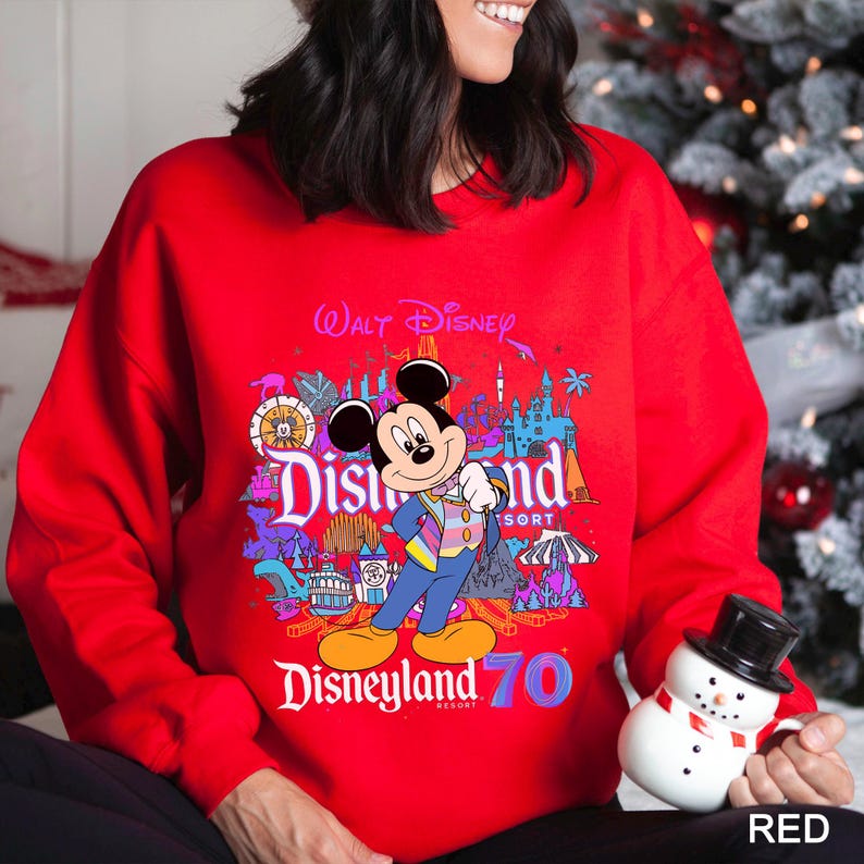 May include: A red sweatshirt featuring a colorful graphic of Mickey Mouse in front of Disneyland Resort imagery. The design includes the text "Walt Disney" and "Disneyland 70". The sweatshirt is a vibrant red color.