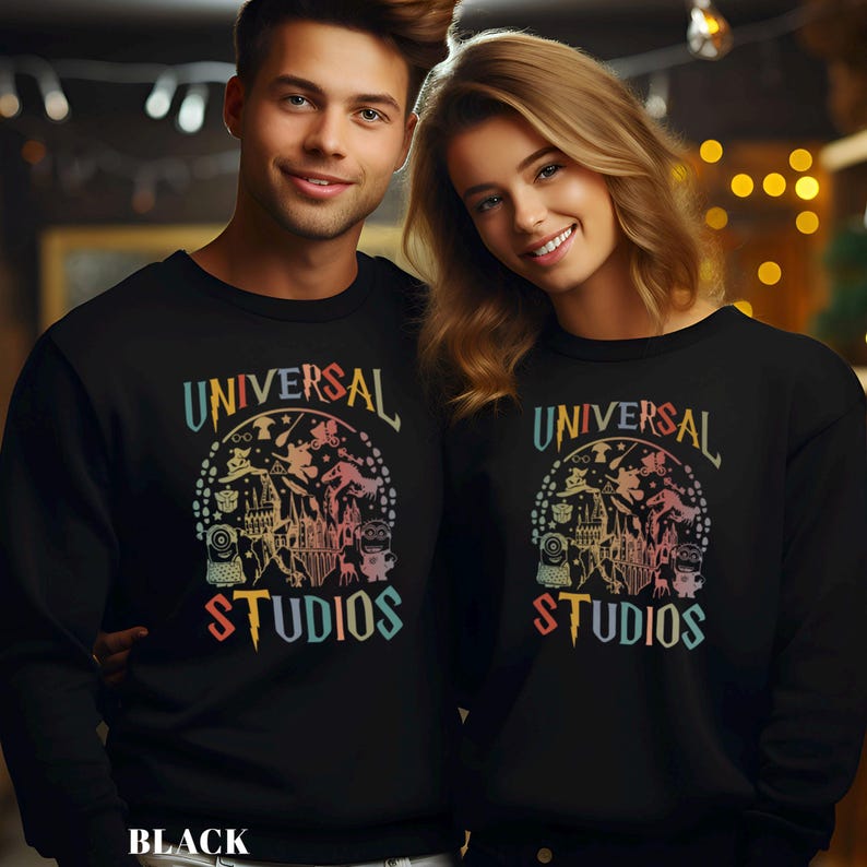 Pode incluir: Duas pessoas vestindo moletons pretos com o texto "UNIVERSAL STUDIOS" em cores de arco-&iacute;ris. O design apresenta personagens de desenhos animados e um castelo. A palavra "BLACK" est&aacute; na parte inferior.
