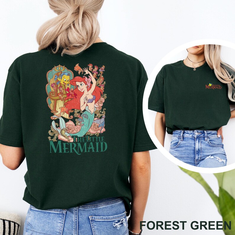 May include: A forest green t-shirt featuring a large graphic on the back of Ariel and Flounder from The Little Mermaid surrounded by floral elements and the text The Little Mermaid. The front chest area displays a small The Little Mermaid logo.
