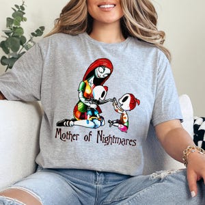 Puede incluir: Camiseta gris con un gr&aacute;fico colorido de un personaje con cabello rojo y un vestido de retazos, sosteniendo a un ni&ntilde;o esqueleto, con otro ni&ntilde;o cerca. El texto "Mother of Nightmares" est&aacute; debajo.