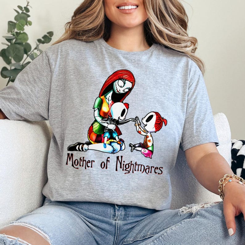 Puede incluir: Camiseta gris con un gr&aacute;fico colorido de un personaje con cabello rojo y un vestido de retazos, sosteniendo a un ni&ntilde;o esqueleto, con otro ni&ntilde;o cerca. El texto "Mother of Nightmares" est&aacute; debajo.