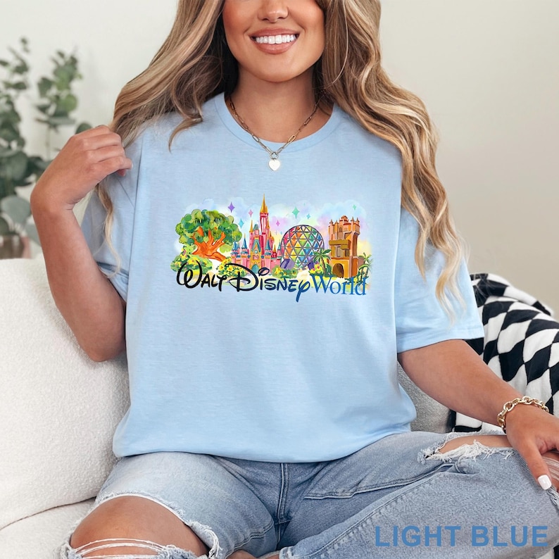 May include: A light blue t-shirt featuring a colorful graphic print of the Walt Disney World theme park landmarks, including the Tree of Life, Cinderella Castle, Spaceship Earth, and the Twilight Zone Tower of Terror, with the text Walt Disney World.