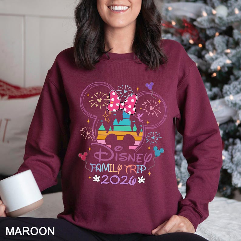 May include: Maroon sweatshirt with a colorful Disney Family Trip 2026 graphic. The design features a Minnie Mouse bow, fireworks, and a castle silhouette. The sweatshirt is a solid maroon color.