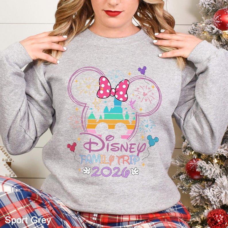 May include: Sport Grey sweatshirt featuring a Disney Family Trip 2026 design. The design includes a castle, a pink polka dot bow, and fireworks within a Mickey Mouse head outline. The text "Disney Family Trip 2026" is in a colorful font.