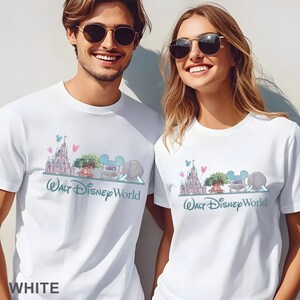 Op de afbeelding: Twee witte t-shirts met een aquarel-stijl afbeelding van de Walt Disney World skyline, inclusief het kasteel, de boom, het studio-bord en de geodetische bol, met de tekst Walt Disney World in een blauwgroen scriptlettertype onder de iconen.