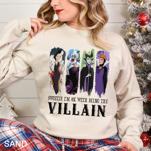 Puede incluir: Una sudadera de color arena con cuatro personajes de villanos de dibujos animados en paneles verticales. El texto "Sweetie I'm OK with being the Villain" est&aacute; impreso debajo de los personajes en negrita negra. La persona lleva un pantal&oacute;n de pijama a cuadros.