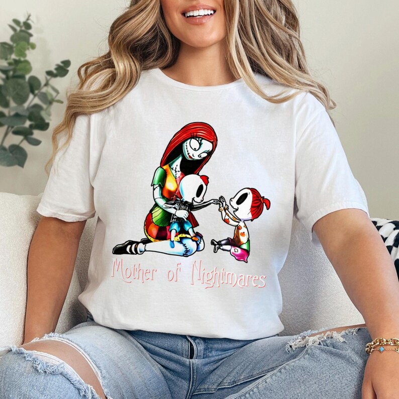 Puede incluir: Camiseta blanca con una ilustraci&oacute;n colorida de un personaje con cabello rojo y una cara cosida, sosteniendo dos figuras esquel&eacute;ticas. El texto "Mother of Nightmares" est&aacute; impreso en rosa debajo de la ilustraci&oacute;n. La camiseta es una prenda casual.