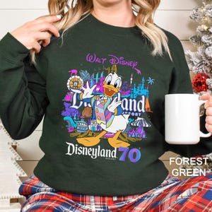 May include: Forest green sweatshirt featuring a colorful graphic of Donald Duck in front of Disneyland Resort attractions. The text "Walt Disney Disneyland Resort 70" is displayed in a vibrant, multi-colored font. A white mug is held in the hand.