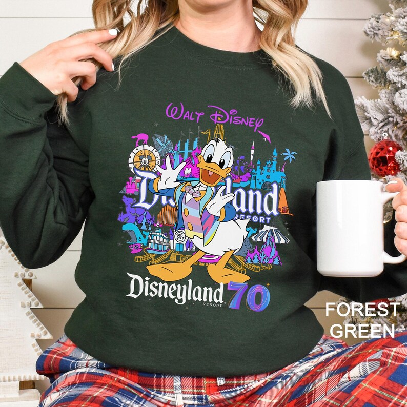 May include: Forest green sweatshirt featuring a colorful graphic of Donald Duck in front of Disneyland Resort attractions. The text "Walt Disney Disneyland Resort 70" is displayed in a vibrant, multi-colored font. A white mug is held in the hand.