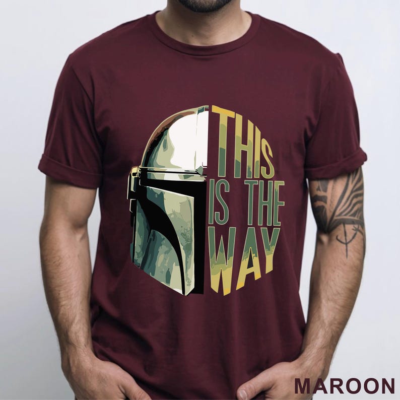 Pu&ograve; includere: Una t-shirt bordeaux a maniche corte con una stampa grafica di un casco metallico sul lato sinistro e il testo "THIS IS THE WAY" in lettere gialle e verdi sul lato destro. Il design presenta una sovrapposizione con motivo mimetico.
