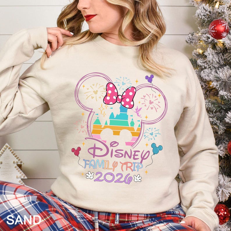 May include: A sand-colored sweatshirt featuring a colorful Disney castle design within a Mickey Mouse head outline. The design includes a pink bow, fireworks, and the text "Disney Family Trip 2026".