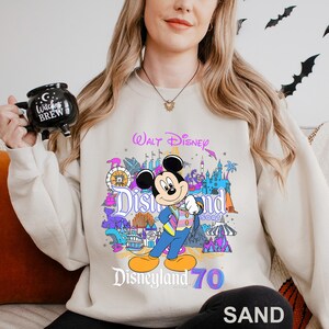 May include: Sand-colored sweatshirt featuring a colorful graphic of Mickey Mouse in front of Disneyland Resort. The design includes the words "Walt Disney" and "Disneyland 70". The sweatshirt is a crew neck style.
