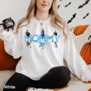 K&ouml;nnte beinhalten: Ein wei&szlig;es Langarm-Sweatshirt mit dem Wort MOMMY in einer blauen, eisigen Schriftart, umgeben von aufgedruckten Charakteren aus dem Film Die Eisk&ouml;nigin, darunter Elsa, Anna, Kristoff, Sven, Olaf und Marshmallow, mit einer schwarzen Witches Brew Tasse.