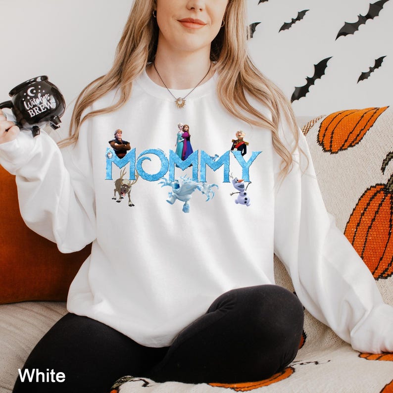 K&ouml;nnte beinhalten: Ein wei&szlig;es Langarm-Sweatshirt mit dem Wort MOMMY in einer blauen, eisigen Schriftart, umgeben von aufgedruckten Charakteren aus dem Film Die Eisk&ouml;nigin, darunter Elsa, Anna, Kristoff, Sven, Olaf und Marshmallow, mit einer schwarzen Witches Brew Tasse.