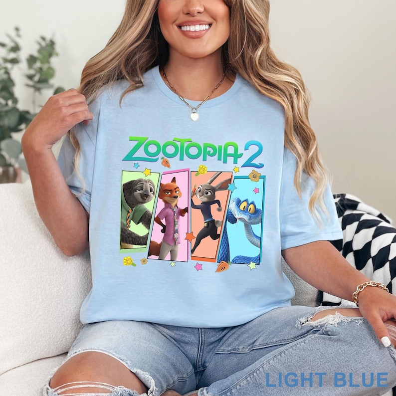 May include: Light blue t-shirt featuring the text "ZOOTOPIA 2" above four character panels. The panels display cartoon images of a sloth, fox, rabbit, and snake. The shirt is light blue and the words "LIGHT BLUE" are printed at the bottom.